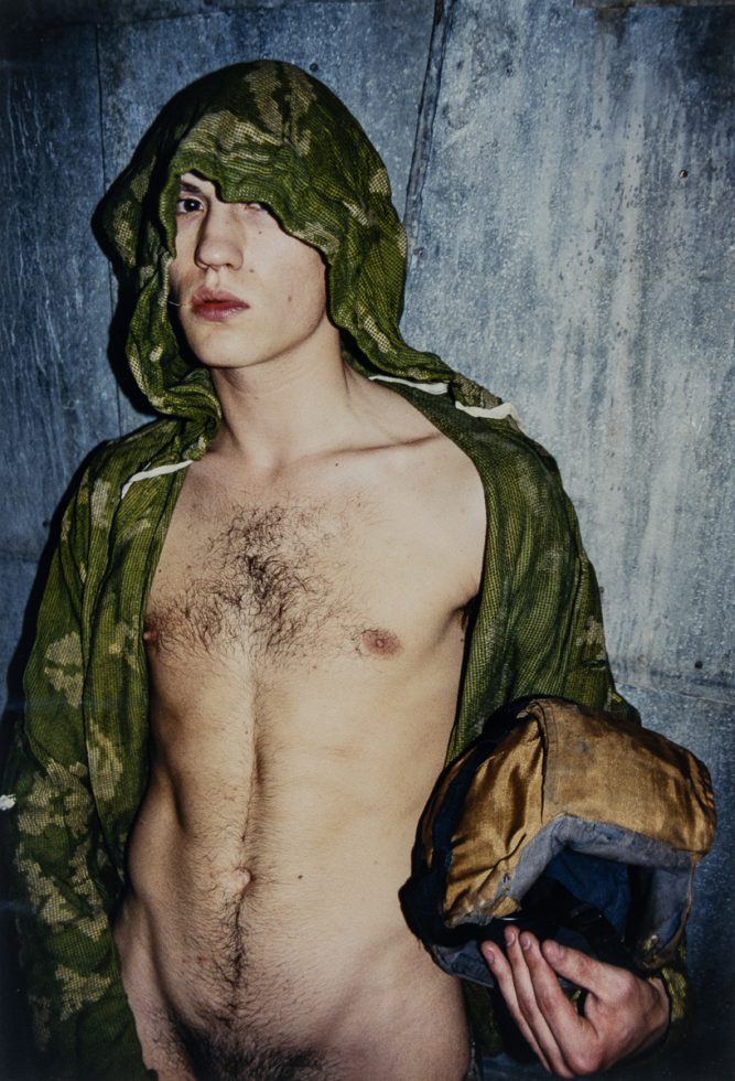 This is a color photograph of a nude young man holding a helmet wearing an army green windbreaker over his shoulders and head.