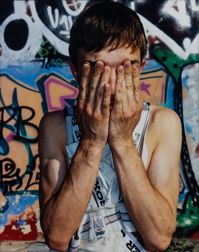 This is a color photograph of a boy in a tank top standing in front of graffiti covering his eyes with his hands.