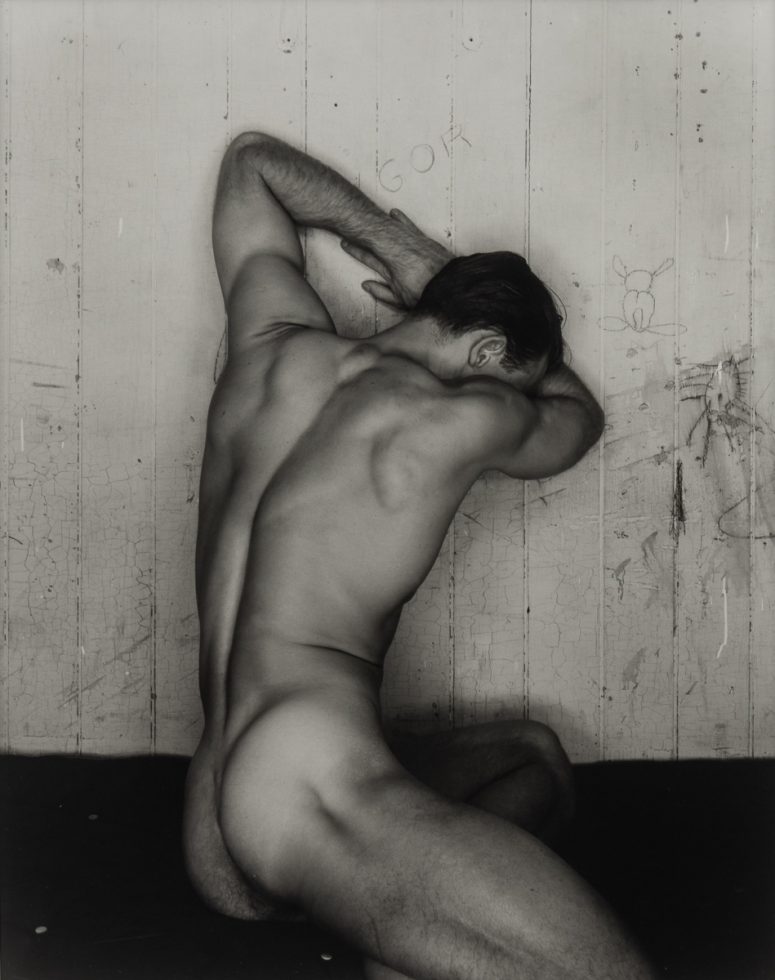 This is a black-and-white photograph of a nude man seated and leaning on a wall with his face away from the camera.