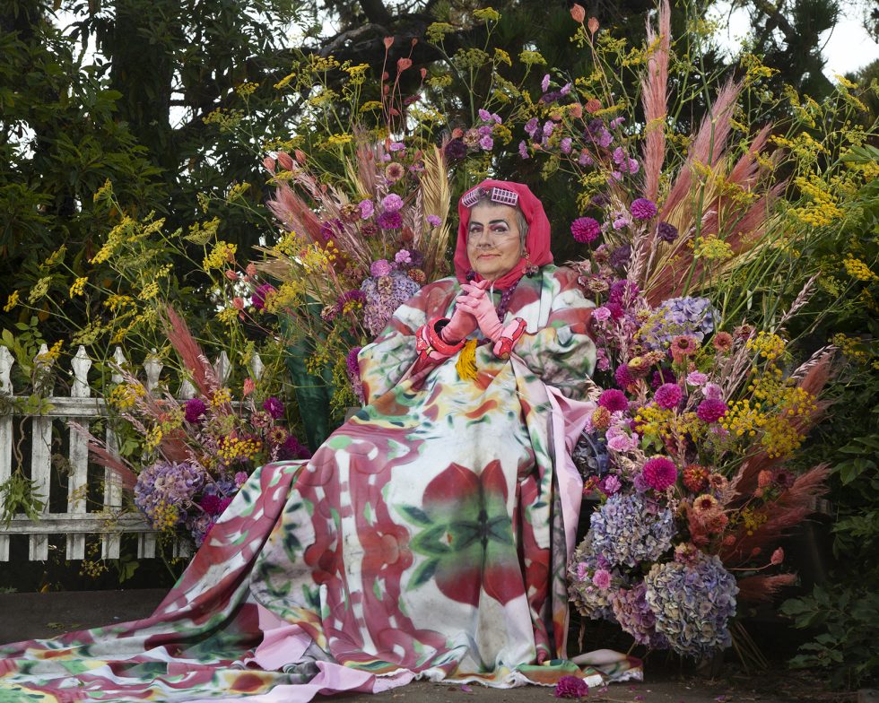 This is a color photograph of an elderly drag performer seated in front of a flower arrangement.