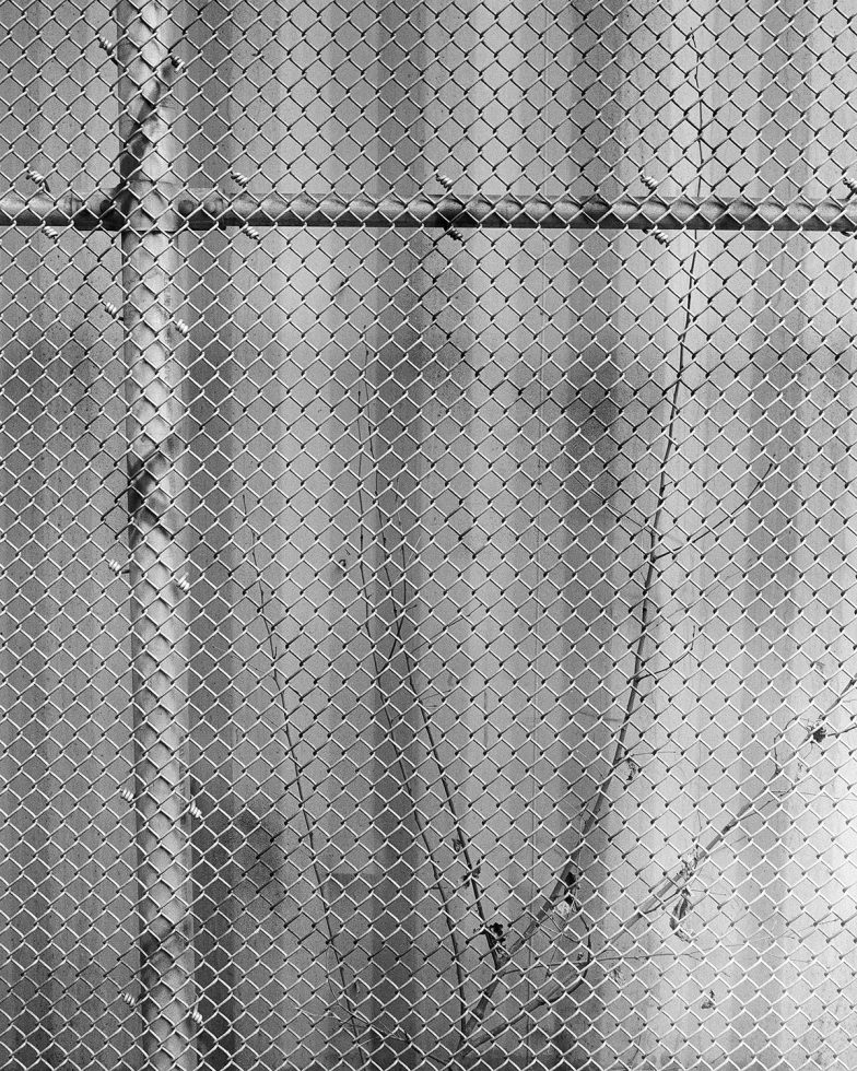 This is a black and white photograph of a shipping container taken through a metal fence.