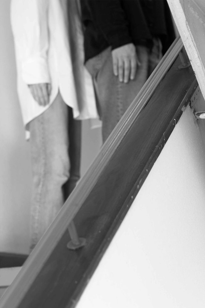 This is a black and white photograph of two people standing with their arms down next to a staircase.