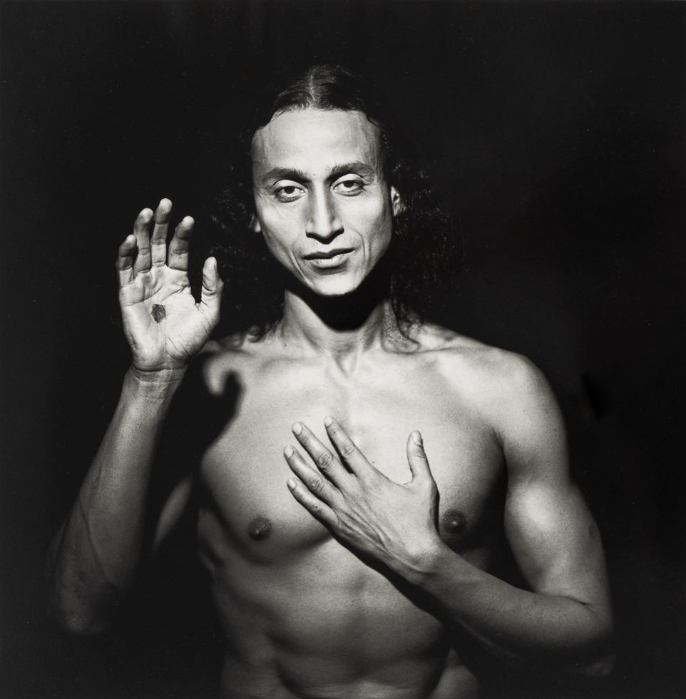 This is a black and white photograph of a shirtless man with one hand on his chest and the other held up with an open palm.