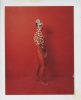 This is a photograph of a nude man in red body paint against a red backdrop with white flower petals decorating his face and body.