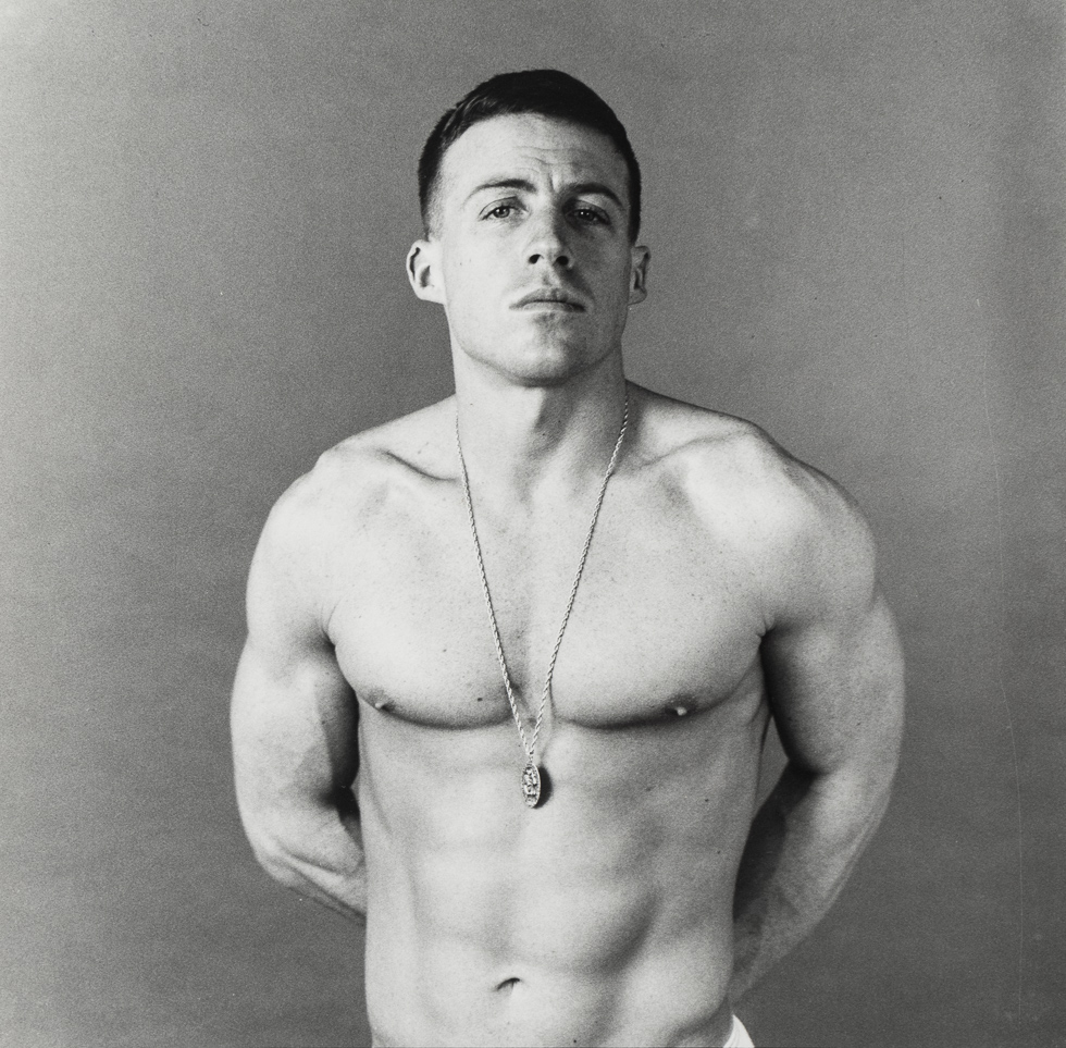 This is a black and white photograph of a shirtless handsome man wearing a necklace with his hands behind his back.