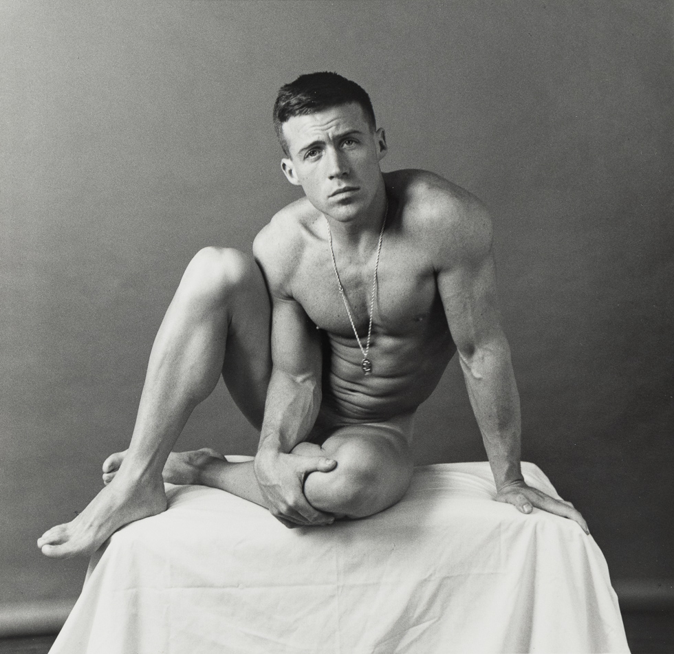This is a black and white photograph of a nude handsome man sitting on a cloth-covered table.