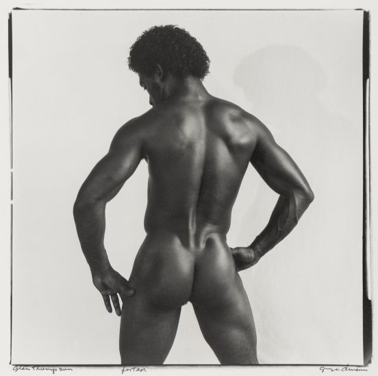 Click for sizes / editions / prices. Black and white photograph of a nude male model, seen from behind, with a muscular physique. The model stands with hands on hips, showcasing his defined back, arms, and glutes. The lighting highlights the contours of his muscles. The photograph appears to be a professional studio portrait.