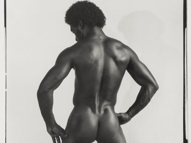 Click for sizes / editions / prices. Black and white photograph of a nude male model, seen from behind, with a muscular physique. The model stands with hands on hips, showcasing his defined back, arms, and glutes. The lighting highlights the contours of his muscles. The photograph appears to be a professional studio portrait.