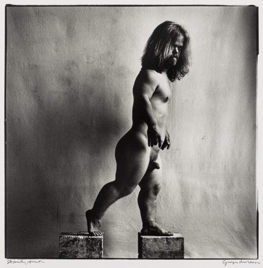 Click for sizes / editions / prices. A black and white photograph of a nude man with dwarfism, standing in profile on two blocks. The man's left foot is on the lower block, and his right foot is on the higher block, as if stepping upward. He has long hair and a beard, and his gaze is directed downward. The lighting and shadows accentuate the contours of his body. The photograph has the artist's signature, "George Dureau," on the bottom right corner and another signature, "Stanley Hurd," on the bottom left corner.