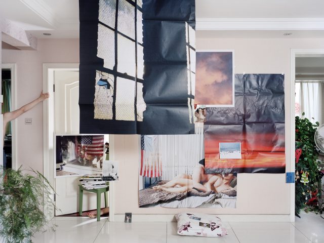 The image shows a room with various large photographs and posters taped to the walls. The images on the posters include scenes of windows, American flags, a sky with clouds, a beach, and a couple lying together. A green stool with books and smaller photographs is in front of the wall. There is a houseplant on the left side and another room with a fan and more plants visible through an open door on the right. A person's arm is visible on the left, partially obscured by a wall, and another person's feet are visible peeking from behind the stool. A pillow with butterfly prints is on the floor in the foreground.