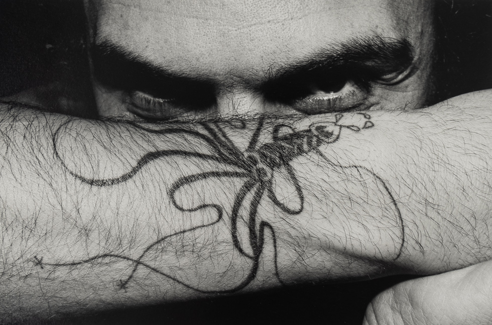 A black and white photograph of a man's eyes and nose, with his forearm covering his nose and highlighting his eyes, with a squid tattoo.