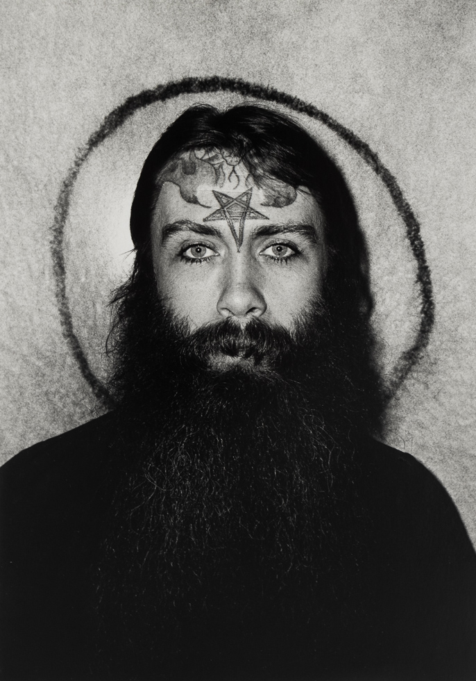 A black and white photograph of a man in a black shirt with a big black beard and a star tattooed on his forehead.