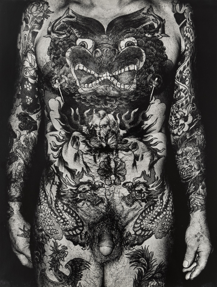 A black and white photograph of a man from the neck down completely covered in intricate tattoos.