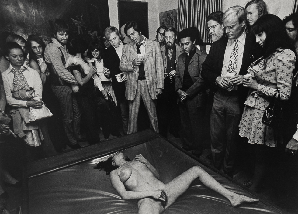 A black and white photograph of a nude woman masturbating on the floor with a crowd observing.