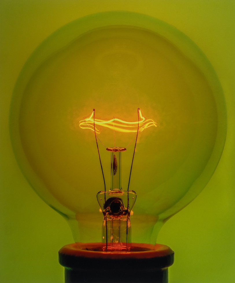 Close-up of a glowing light bulb filament against a vivid green background. The orange-yellow light curves intricately, highlighting energy and warmth.