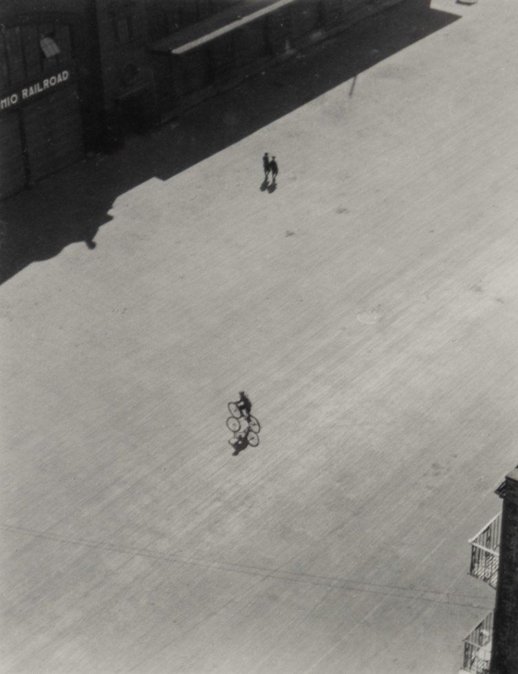 A high-angle black and white photo shows a lone cyclist and two figures on horseback in a large, empty paved area with a building in the background.
