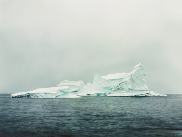A jagged, white iceberg floats in calm, dark water. The iceberg's peaks rise sharply against a pale gray sky