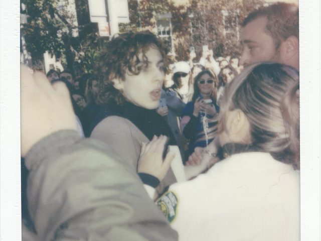 Click for sizes / editions / prices. A polaroid of a group of people in a crowd with a young man with curly dark hair in the center of the scene.