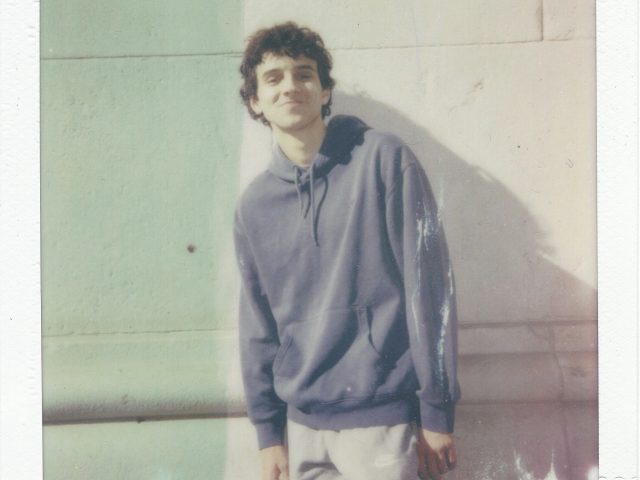 Click for sizes / editions / prices. A polaroid photograph of a young man in a grey hoodie against a stone wall.