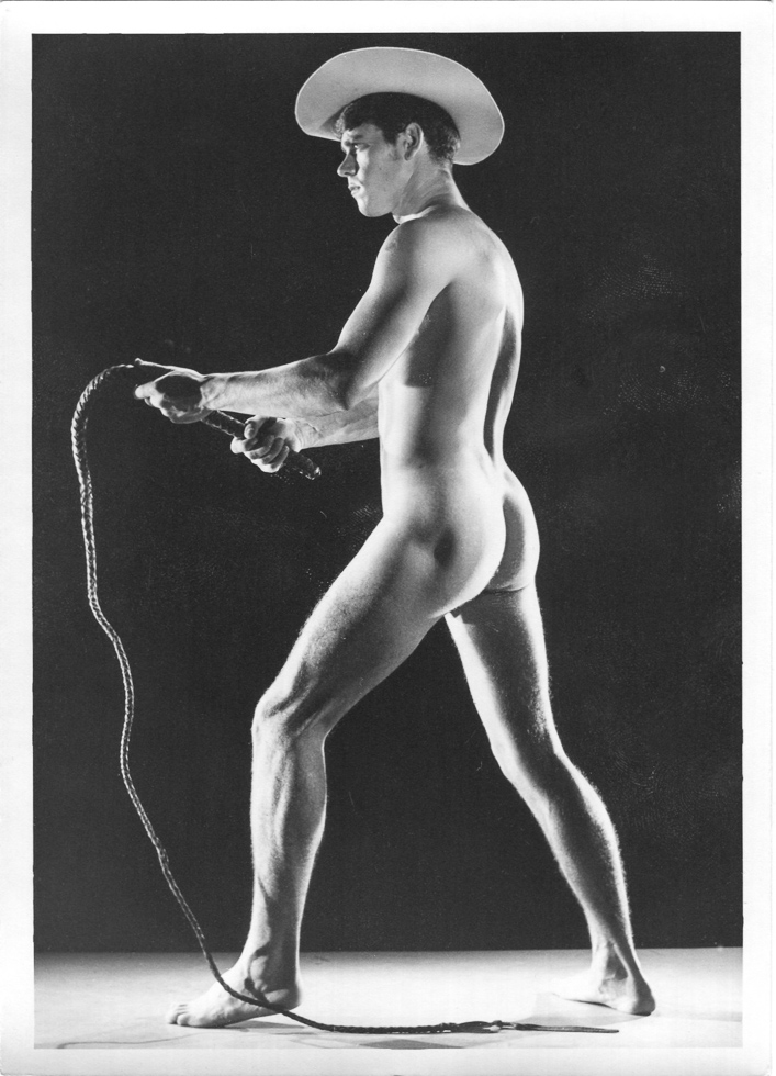 A black and white photograph of a nude man wearing a cowboy hat holding a whip.