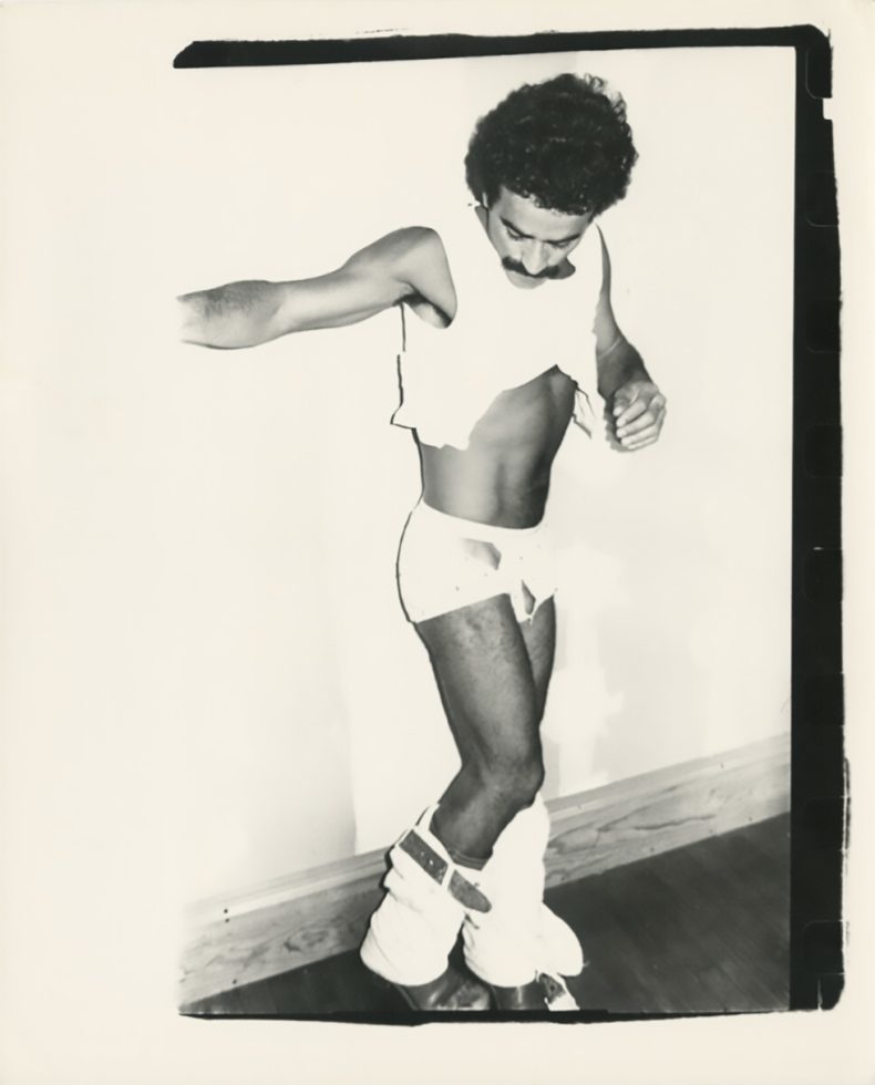 A black and white photograph of a man with dark curly hair in short white shorts.