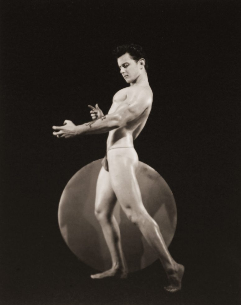 A black and white photograph of a muscular man standing next to a circle in front of a black background.