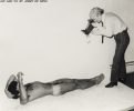 A black and white photograph of a nude man lying on the ground with a man standing above him with a camera.