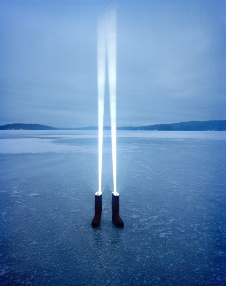 Black boots on an icy surface emit bright vertical beams of light into a pale blue sky, creating a surreal and serene winter landscape.