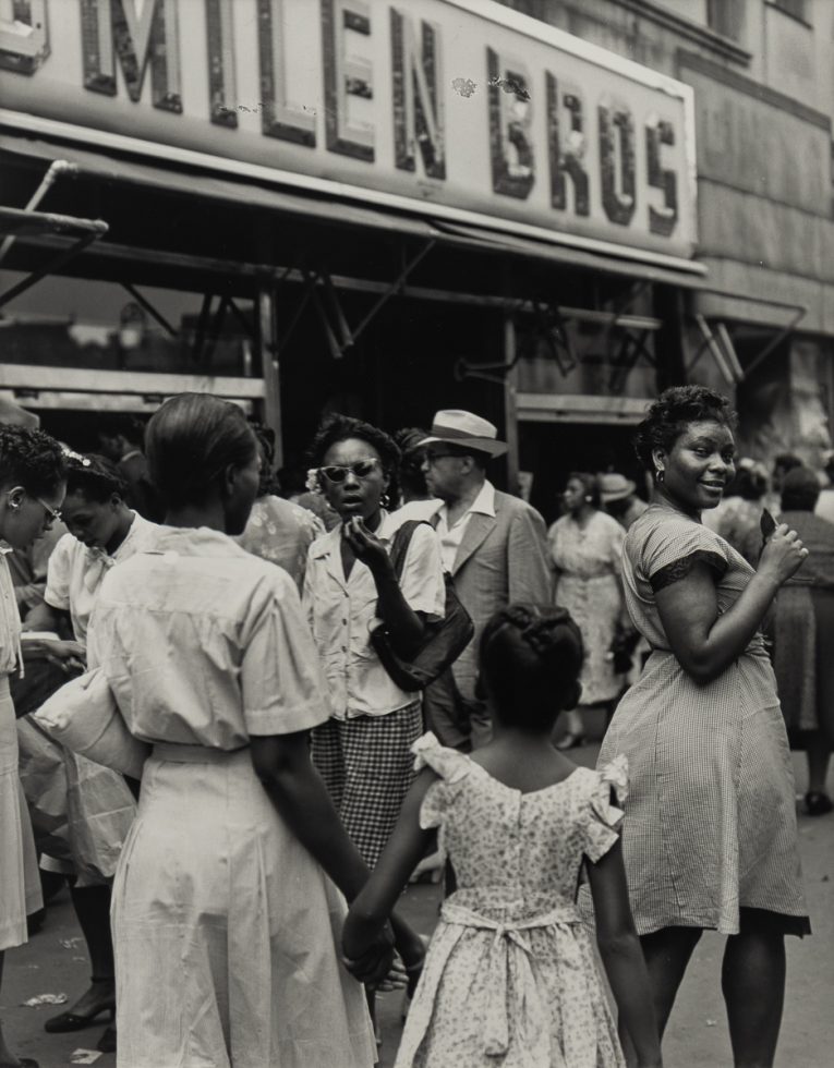 A lively street scene in Harlem, New York, featuring a crowd of mostly Black individuals in front of a "Smilen Bros" storefront. A woman in a checkered dress looks back, smiling.