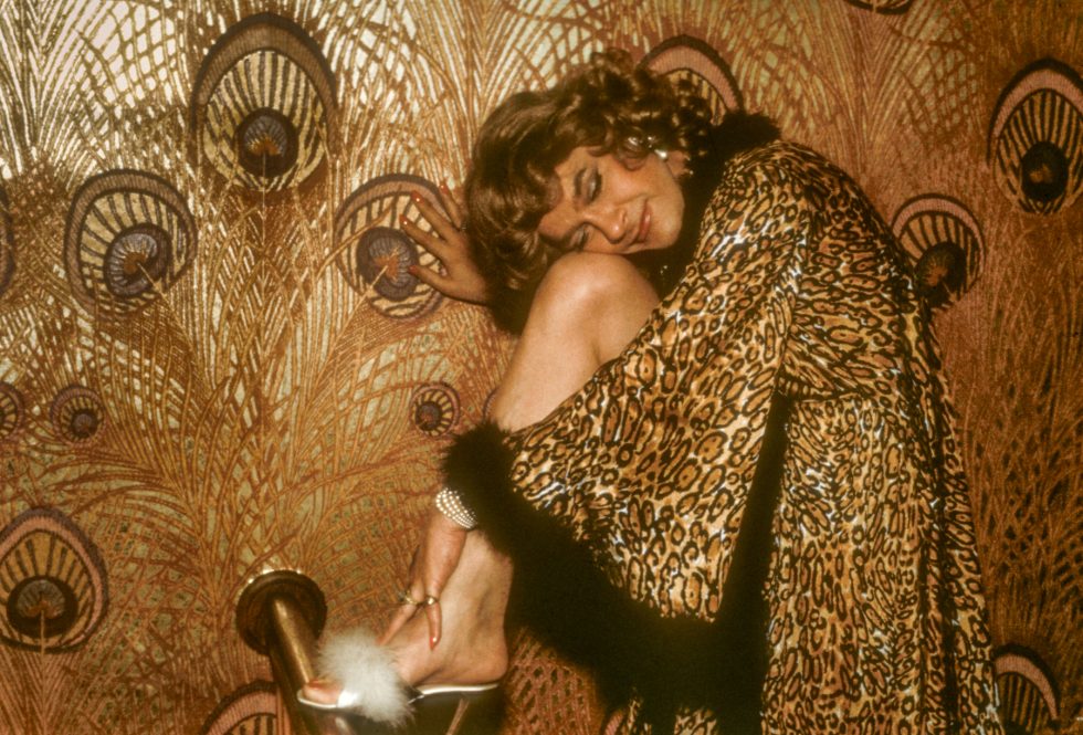 A person in a leopard-print robe and fluffy heels leans against a gold peacock-patterned wall, smiling with eyes closed.