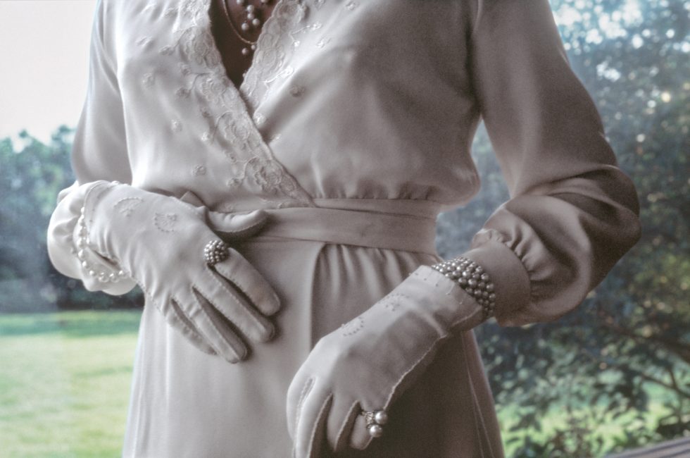 A person wearing an elegant white gown with lace details, pearl-adorned gloves, and jewelry, posing with hands gently placed on their waist.
