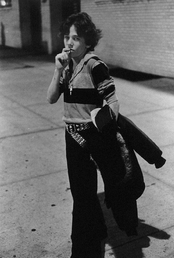 A black-and-white photograph of a young person with curly hair standing on a city sidewalk at night.