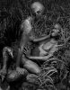 Two nude figures covered in mud embrace in a grassy, wetland environment, gazing intensely at each other.