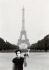 A black-and-white photograph of a man standing near the Eiffel Tower holding a burning object with a historical portrait of a young man superimposed on his face.