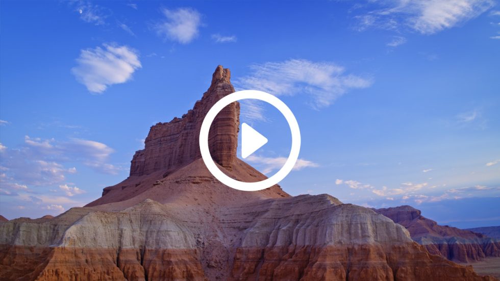 A color image of massive sandstone formations against a blue sky with scattered little white clouds.