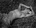 A shirtless person with short hair and a pierced navel lies in a grassy field with their arm resting over their head, holding a daisy near their face.