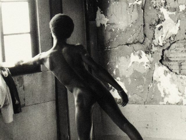 A nude figure stands in a ballet pose near a window in a decaying room with peeling walls and debris on the floor.