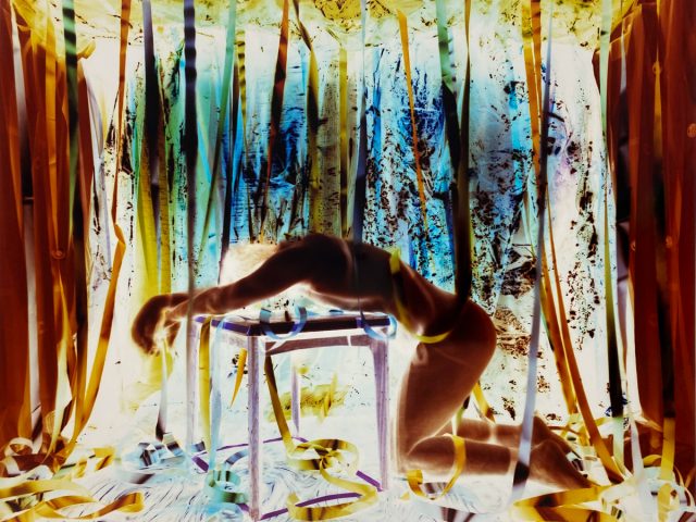 A nude, blurred figure kneels with arms draped over a table, surrounded by hanging ribbons in a surreal, high-contrast environment.