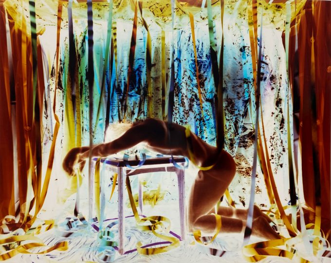 A nude, blurred figure kneels with arms draped over a table, surrounded by hanging ribbons in a surreal, high-contrast environment.