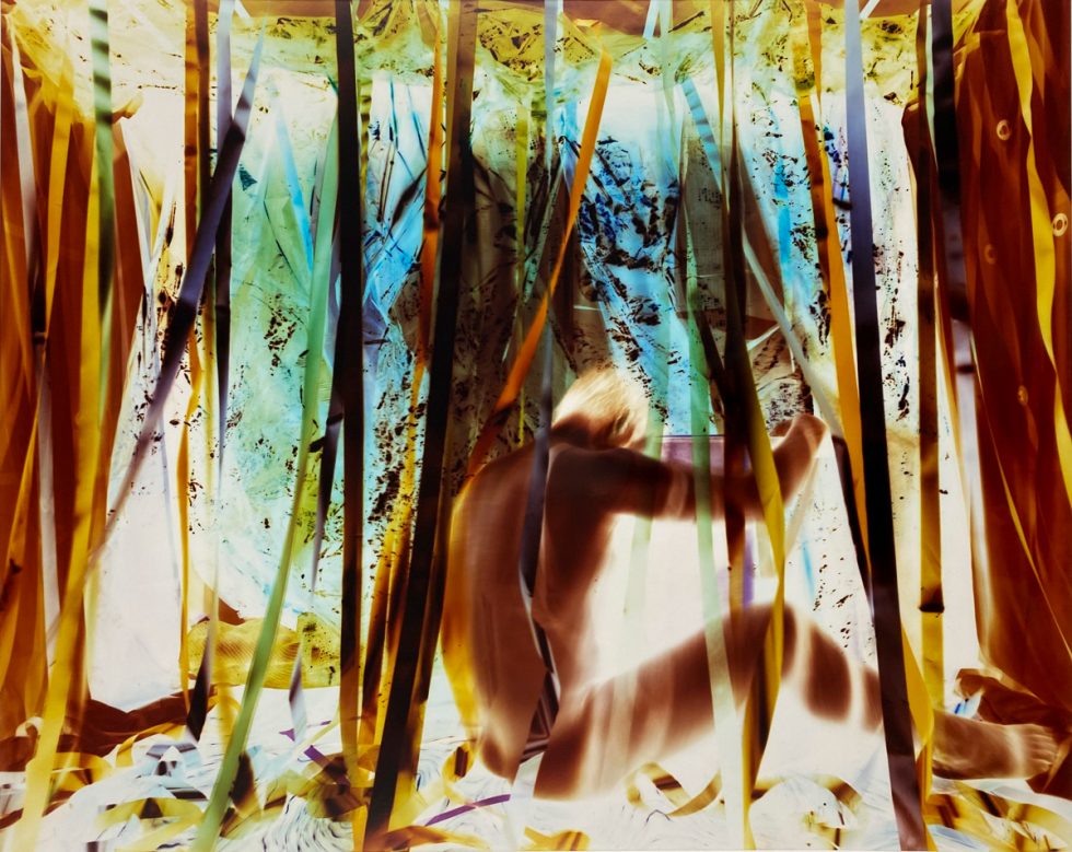 A blurred figure sits among colorful, vertical ribbons in a vivid, abstract setting with a surreal, negative-like color palette.