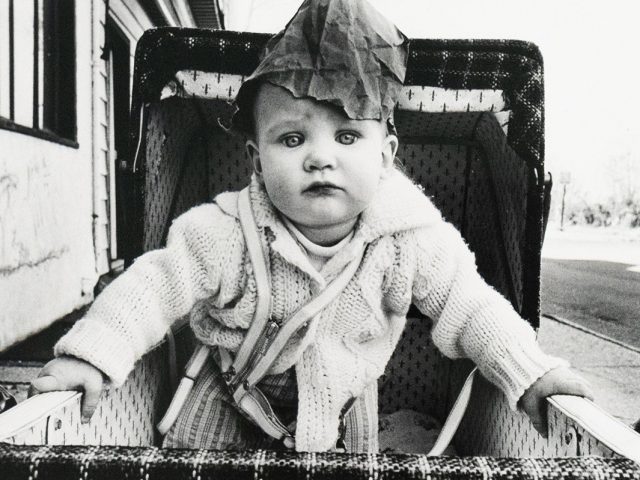 A black and white photograph of a baby in a stroller wearing a paper bag on its head while staring into the camera.