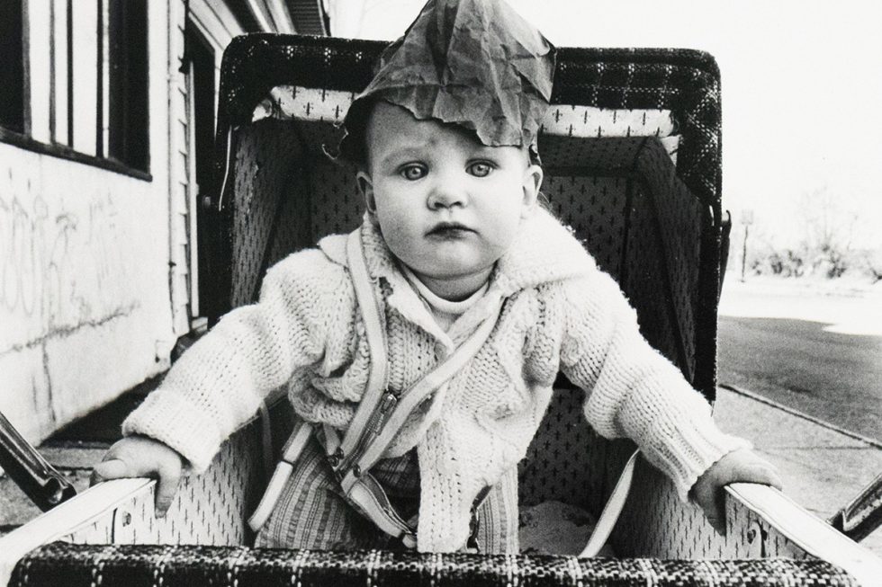 A monochrome photograph captures a baby in a stroller, wearing a light-colored sweater, suspenders, and a dark, crumpled hat, looking directly at the camera. The setting includes a building to the left and an outdoor area to the right.