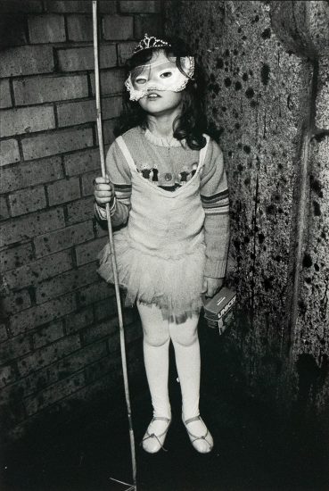 A young child wearing a decorative mask and a tutu stands holding a thin pole in one hand and a small box labeled "UNICEF" in the other. The child is positioned against a textured wall that transitions from brick on the left to a darker, uneven surface on the right.