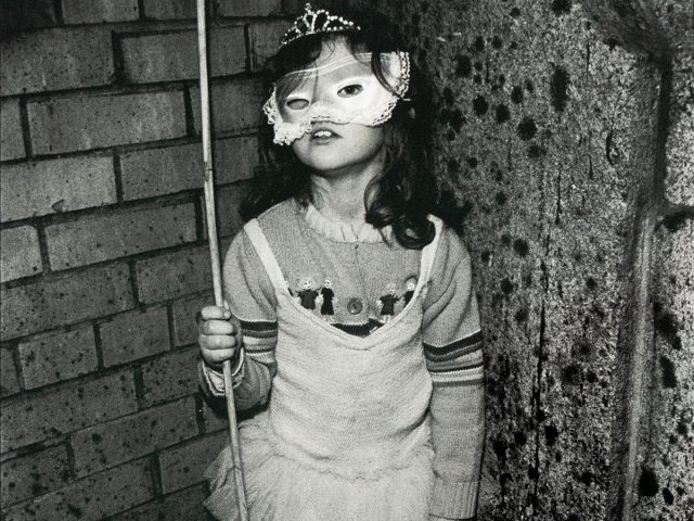 A young child wearing a decorative mask and a tutu stands holding a thin pole in one hand and a small box labeled "UNICEF" in the other. The child is positioned against a textured wall that transitions from brick on the left to a darker, uneven surface on the right.