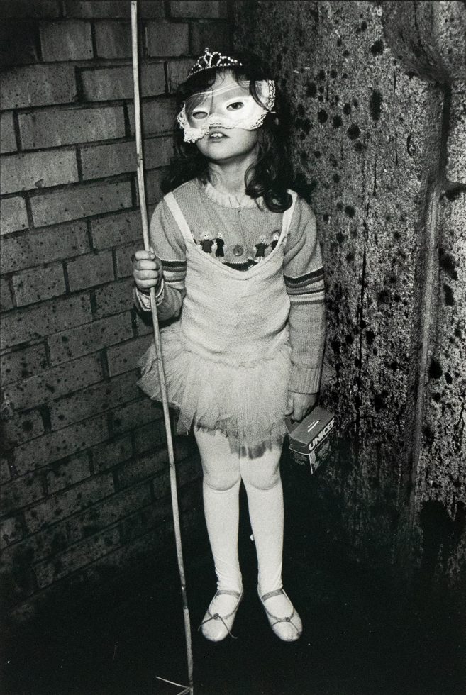 A young child wearing a decorative mask and a tutu stands holding a thin pole in one hand and a small box labeled "UNICEF" in the other. The child is positioned against a textured wall that transitions from brick on the left to a darker, uneven surface on the right.