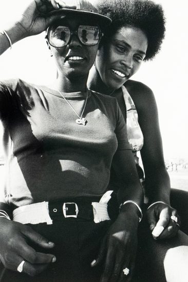 Two Black women are closely posed in a black and white photograph, smiling towards the camera. The woman in the foreground wears sunglasses, a cap, a t-shirt, and a belt, while the woman behind has an afro and a patterned top.