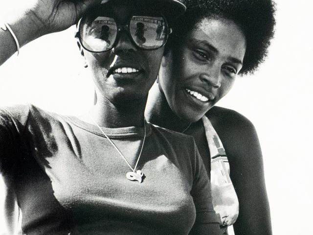 Two Black women are closely posed in a black and white photograph, smiling towards the camera. The woman in the foreground wears sunglasses, a cap, a t-shirt, and a belt, while the woman behind has an afro and a patterned top.