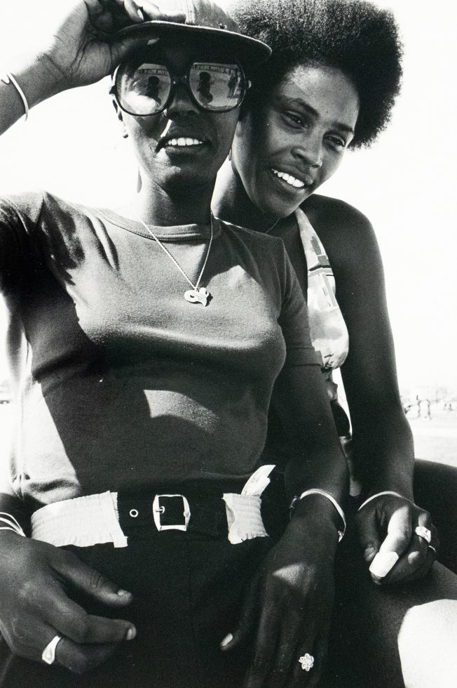 Two Black women are closely posed in a black and white photograph, smiling towards the camera. The woman in the foreground wears sunglasses, a cap, a t-shirt, and a belt, while the woman behind has an afro and a patterned top.