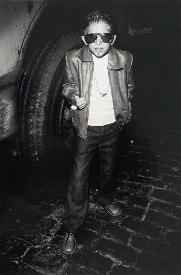A boy wearing sunglasses, a leather jacket, and jeans stands on a brick surface, holding a cigarette in his mouth and giving a thumbs up.
