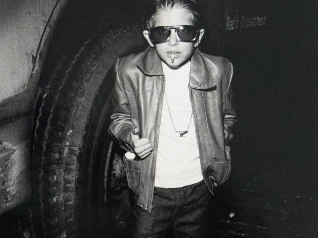 A boy wearing sunglasses, a leather jacket, and jeans stands on a brick surface, holding a cigarette in his mouth and giving a thumbs up.